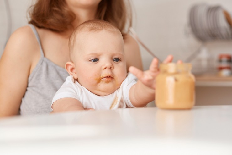 curious-child-stretching-out-arm-feed-jar-faceless-mother-feeds-her-little-infant-daughter-with-vegetable-puree-sitting-table-home-feeding-up